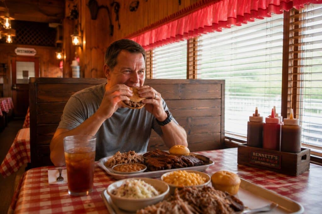 Family enjoying a barbecue dinner in a rustic Gatlinburg restaurant while a husband eats a pulled pork sandwich during a Smoky Mountain vacation.
