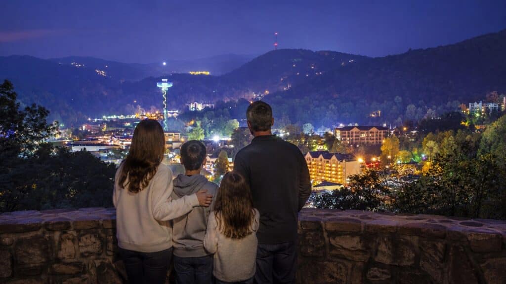 Family of four standing at a stone overlook wall above downtown Gatlinburg at night with the Smoky Mountains fading into blue twilight.