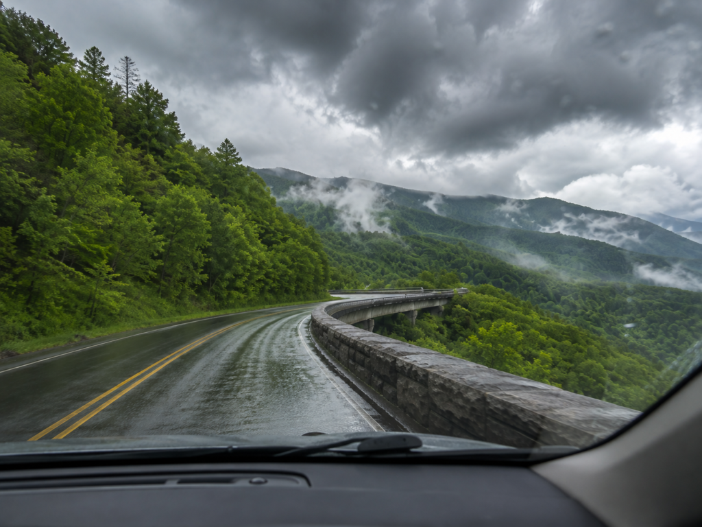 View through a car windshield of wet Foothills Parkway curving through green mountains beneath storm clouds