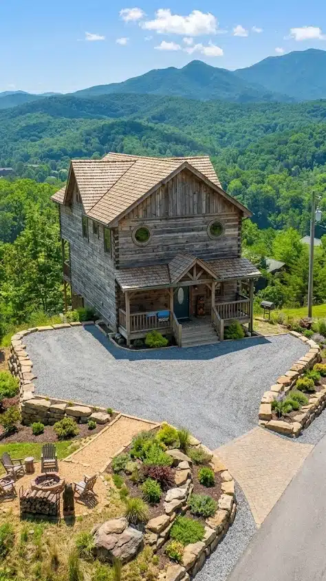Two-story wooden cabin with a curved driveway, landscaped fire pit area, and forested mountains behind it